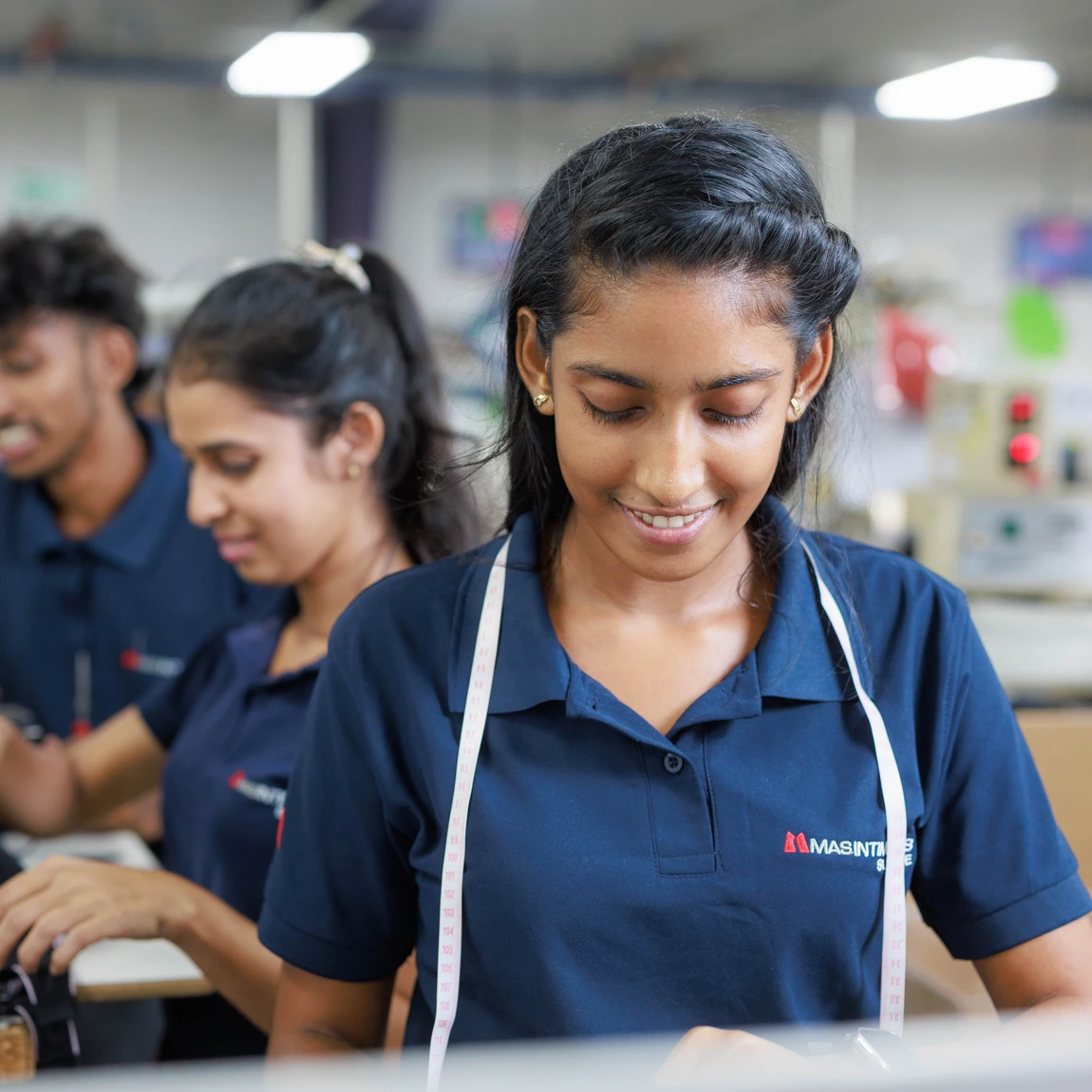 A smiling MAS woman employee with a measuring tape around her neck, with colleagues at sewing stations behind her