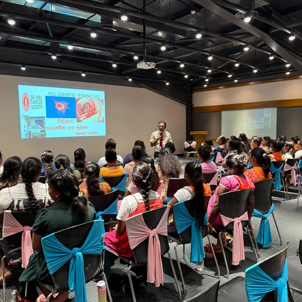 An audience at an MAS gender-based-violence awareness session facing a presenter and a women's-health awareness slide